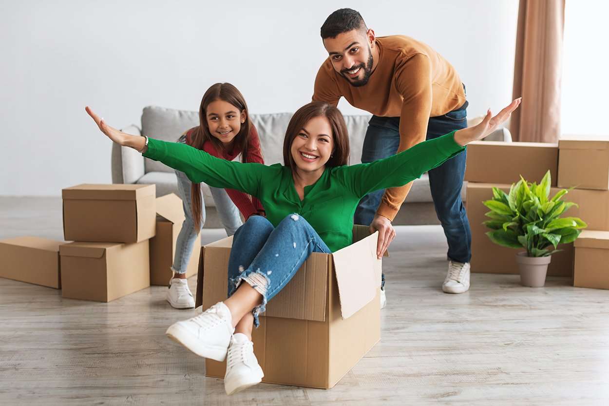 New Home, People And Real Estate Concept. Excited loving family playing having fun celebrating moving day, cheerful Arab man and girl riding happy lady in cardboard box container in living room